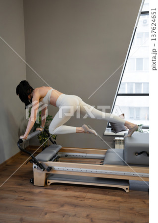 Woman in light gym clothes doing pilates stretches on reformer, knee bent in indoor studio. Pilates stretches, reformer, indoor, fitness, stretch. Woman in light gym clothes doing pilates stretches on reformer, knee bent in indoor studio. Pilates stretches, reformer, indoor, fitness, stretch. 137624651