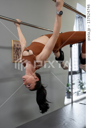 Woman lying back, arching and hanging from pilates bar in gymnasium during advanced workout. Arching, hanging, pilates bar, gymnasium, advanced workout. Woman lying back, arching and hanging from pilates bar in gymnasium during advanced workout. Arching, hanging, pilates bar, gymnasium, advanced workout. 137624746