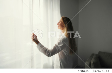 Woman opening window curtains at home in morning Woman opening window curtains at home in morning 137626043