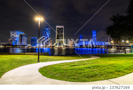 Vibrant night view of downtown Jacksonville, Florida, showcasing the illuminated city skyline, Main Street Bridge, and reflections on the Saint Johns River with a scenic park path Vibrant night view of downtown Jacksonville, Florida, showcasing the illuminated city skyline, Main Street Bridge, and reflections on the Saint Johns River with a scenic park path 137627169