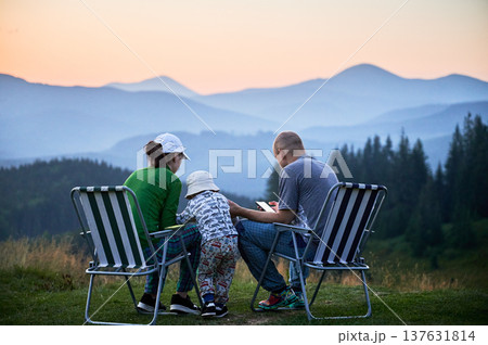 Family of three sits on foldable chairs, enjoying peaceful sunset in mountains, share time together in nature. Child stands between parents. Layered mountain ranges and soft, colorful sky. Family of three sits on foldable chairs, enjoying peaceful sunset in mountains, share time together in nature. Child stands between parents. Layered mountain ranges and soft, colorful sky. 137631814