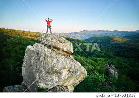 Aerial view of climber standing atop rock formation at Dovbush Rocks in Carpathian mountains, Ukraine. Sun sets, casting warm glow over lush, green forest, distant hills, creating breathtaking view. Aerial view of climber standing atop rock formation at Dovbush Rocks in Carpathian mountains, Ukraine. Sun sets, casting warm glow over lush, green forest, distant hills, creating breathtaking view. 137631815