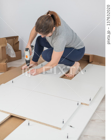 A Caucasian woman uses a screwdriver to tighten a screw while assembling a cabinet. 137632020