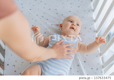 Mother sitting beside the crib gently stroking her baby lying in a cozy bed at home. Warm intimate moment filled with love, trust and calm. Motherhood, infant comfort and emotional bonding concept Mother sitting beside the crib gently stroking her baby lying in a cozy bed at home. Warm intimate moment filled with love, trust and calm. Motherhood, infant comfort and emotional bonding concept 137633475
