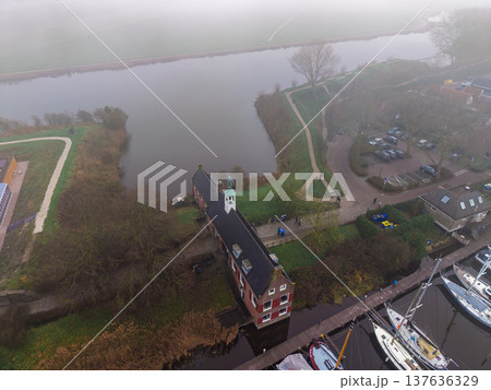 Aerial view of a historic brick building with a white bell tower on a foggy riverbank. Moody morning landscape with a marina, sailboats, and a stone fortress moat in the misty Dutch countryside. Aerial view of a historic brick building with a white bell tower on a foggy riverbank. Moody morning landscape with a marina, sailboats, and a stone fortress moat in the misty Dutch countryside. 137636329