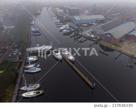 Aerial view of a misty marina with sailboats and yachts docked in calm water. Moody fog over a European harbor with boat warehouses and town houses in the background. Tranquil morning atmosphere. 137636330