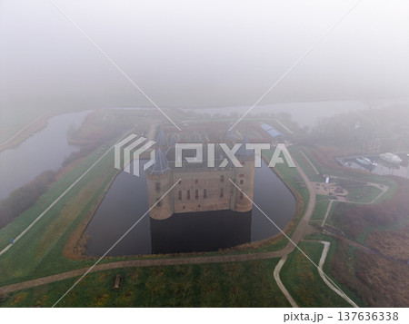 Aerial shot of medieval Muiderslot Castle shrouded in thick fog. Historic brick fortress with towers surrounded by a water moat and misty gardens in the Netherlands. Moody morning atmosphere. Aerial shot of medieval Muiderslot Castle shrouded in thick fog. Historic brick fortress with towers surrounded by a water moat and misty gardens in the Netherlands. Moody morning atmosphere. 137636338