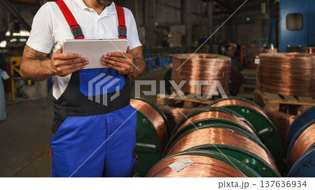 Worker checks data while standing next to coils of copper wire in a warehouse 137636934