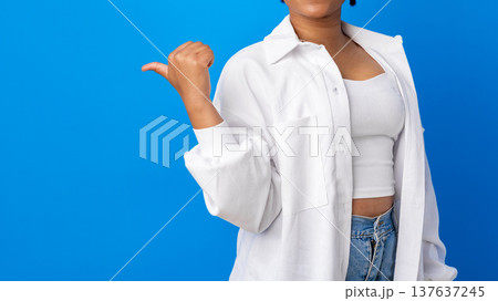 Woman points to the side while standing against a blue background in casual outfit 137637245