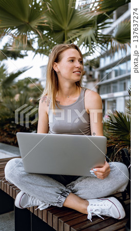 Woman sitting outdoors with a laptop surrounded by plants working on a sunny day 137637274