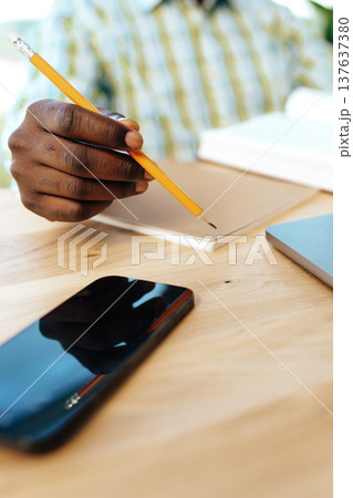 Person writes with a pencil in a notebook while sitting at a wooden table with a phone Person writes with a pencil in a notebook while sitting at a wooden table with a phone 137637380