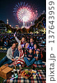 Friends with American flags sitting on rug in park at night watching fireworks display and smiling. Fireworks, friends, night, park, celebration, summer, USA. 137643959