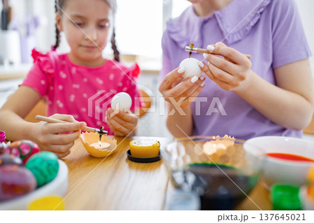 A mother and her daughter are engaged in the traditional activity of decorating Easter eggs with wax and dyes at a wooden table 137645021