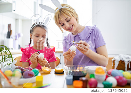 A mother and daughter wearing bunny ears are focused on decorating Easter eggs with wax and paint at a kitchen table filled with colorful eggs and craft supplies A mother and daughter wearing bunny ears are focused on decorating Easter eggs with wax and paint at a kitchen table filled with colorful eggs and craft supplies 137645022