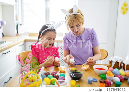 A mother and daughter wearing bunny ears are happily decorating Easter eggs together at a wooden table in a bright kitchen, surrounded by colorful eggs and baking supplies A mother and daughter wearing bunny ears are happily decorating Easter eggs together at a wooden table in a bright kitchen, surrounded by colorful eggs and baking supplies 137645031