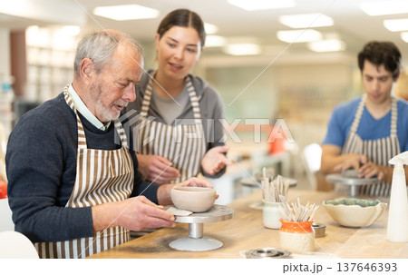 Woman help mature man sculpting ceramic product from raw clay in workshop 137646393