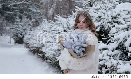 Woman wearing white winter coat and gloves, carrying festive evergreen bouquet while walking through snow covered woodland during holiday season 137646476