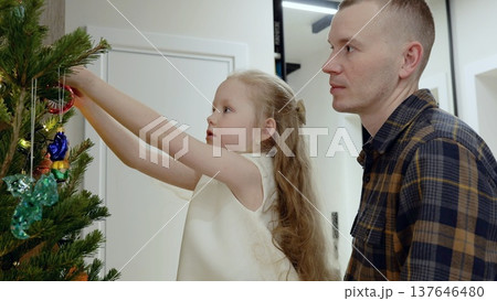 Young girl carefully placing glass ornaments on christmas tree, dad watching, capturing heartwarming holiday bonding moment together Young girl carefully placing glass ornaments on christmas tree, dad watching, capturing heartwarming holiday bonding moment together 137646480