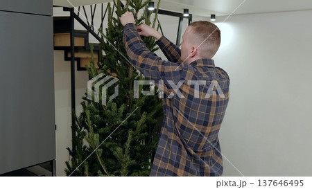 Man decorating contemporary christmas tree, positioning ornaments and branches with precision, creating festive home interior ambiance 137646495