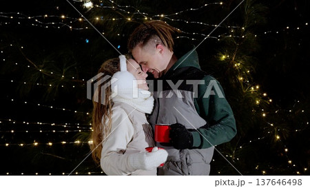 Happy young couple smiling and touching foreheads in front of christmas tree lights, holding red mugs, wearing winter clothes, enjoying christmas time together 137646498