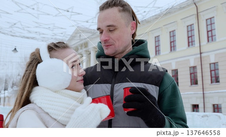 Cozy couple wearing winter attire, holding bright red mugs with steaming drinks, standing together amid snow covered urban scenery radiating holiday warmth Cozy couple wearing winter attire, holding bright red mugs with steaming drinks, standing together amid snow covered urban scenery radiating holiday warmth 137646558
