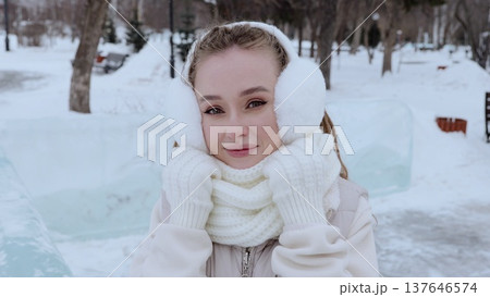 Young woman wearing white earmuffs, gloves, and a scarf, enjoying a winter day in a snowy park, smiling brightly while embracing the chilly weather and serene surroundings 137646574