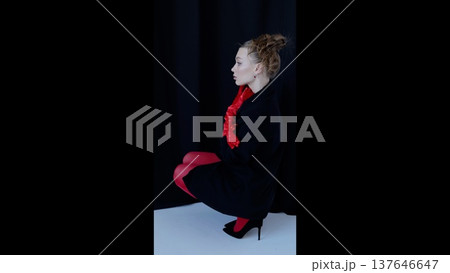 Professional model posing in studio, wearing red tights, gloves, black dress with heels. Dark background emphasizing sleek silhouette and dramatic side profile 137646647