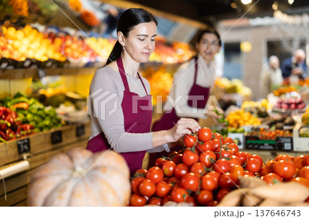 Adult woman seller puts tomatoes in vegetable shop Adult woman seller puts tomatoes in vegetable shop 137646743
