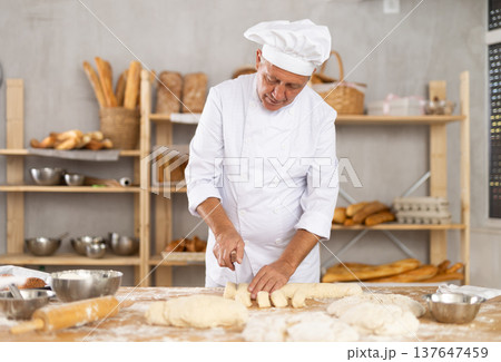 Baker in uniform cutting raw dough with knife to make croissants or buns 137647459