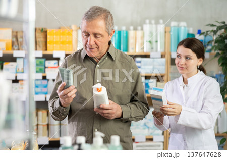 Senior man buys cream at drug store, girl pharmacist in background Senior man buys cream at drug store, girl pharmacist in background 137647628