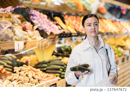 In supermarkets vegetable store, female customer takes some cucumber from display shelf. Woman selects necessary vegetables In supermarkets vegetable store, female customer takes some cucumber from display shelf. Woman selects necessary vegetables 137647672
