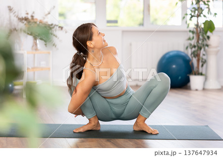 Young woman yoga trainer performing frog pose on black rug Young woman yoga trainer performing frog pose on black rug 137647934