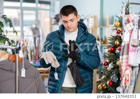 Guy is standing in a store against the background of a Christmas tree and looking at gloves 137648351