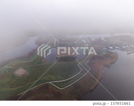 Wide aerial view of Muiderslot Castle in the Netherlands, shrouded in thick morning fog. Medieval fortress with a moat and surrounding star-shaped ramparts in a misty, moody landscape. Wide aerial view of Muiderslot Castle in the Netherlands, shrouded in thick morning fog. Medieval fortress with a moat and surrounding star-shaped ramparts in a misty, moody landscape. 137651661