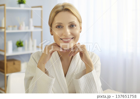 Smiling beautiful woman wearing white robe sitting in beauty clinic room or at home, beauty products Smiling beautiful woman wearing white robe sitting in beauty clinic room or at home, beauty products 137651758