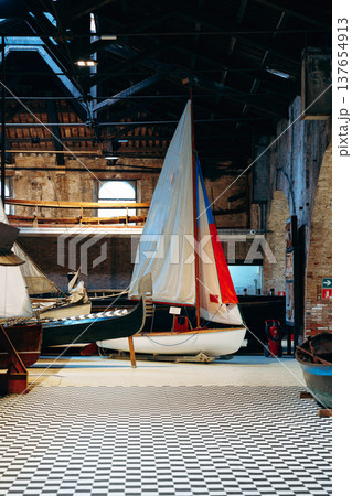 Sailboats and wooden boats inside a large boat house with checkered floor 137654913
