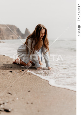 Beach woman waves sitting on sandy shore wearing blue shirt touching ocean foam at sunrise with cliffs in background 137658647