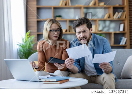 Stressed young couple calculating taxes and analyzing domestic bills, feeling anxiety about financial management and household budgeting while sitting on a sofa with a laptop Stressed young couple calculating taxes and analyzing domestic bills, feeling anxiety about financial management and household budgeting while sitting on a sofa with a laptop 137667354