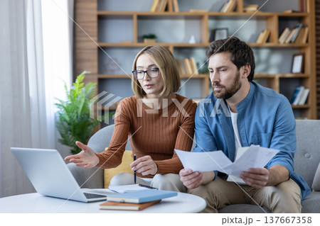 Couple analyzing financial documents and using a laptop for money management, budgeting, and planning expenses together on a sofa in their living room 137667358