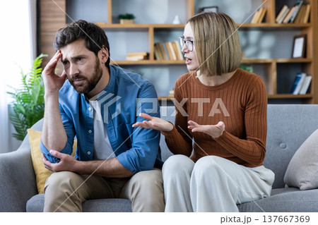 Couple sitting on a couch in a living room having an argument, with the man looking upset and distressed, while the woman expresses her feelings with an open hand gesture 137667369