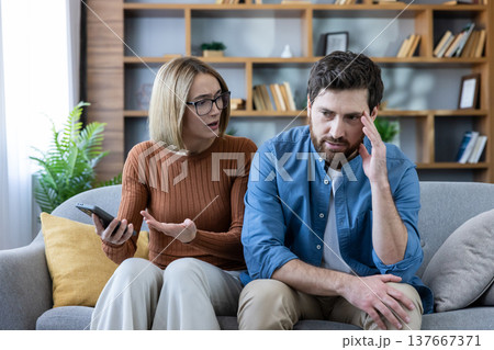 Upset woman holding a smartphone and arguing with frustrated man on sofa, highlighting relationship problems, stress, betrayal, and jealousy in domestic life 137667371