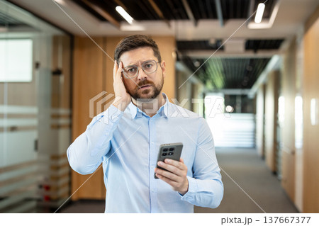 Man in office hallway experiencing stress and frustration, holding a smartphone and reacting to bad news or a difficult message with a confused and worried expression Man in office hallway experiencing stress and frustration, holding a smartphone and reacting to bad news or a difficult message with a confused and worried expression 137667377