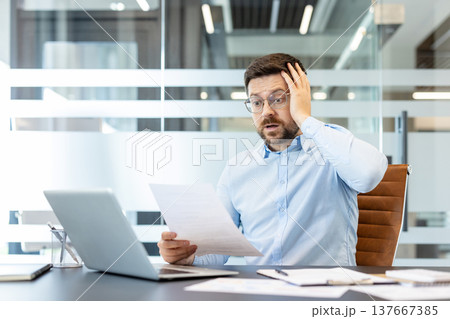 Businessman receiving stressful news while reviewing an official document, reacting with shock and worry about financial problems, debt, or an unforeseen crisis at work 137667385