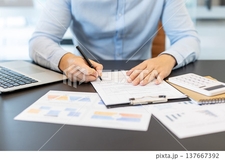 Businessman using a pen to sign a form on a clipboard, reviewing financial documents, graphs, and a calculator on a desk, representing accounting, analysis, and data management Businessman using a pen to sign a form on a clipboard, reviewing financial documents, graphs, and a calculator on a desk, representing accounting, analysis, and data management 137667392
