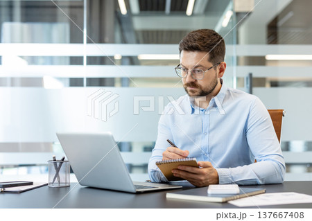 Businessman in glasses and a light blue shirt actively taking notes in a spiral notebook while focusing on his laptop computer, working diligently at a modern office desk Businessman in glasses and a light blue shirt actively taking notes in a spiral notebook while focusing on his laptop computer, working diligently at a modern office desk 137667408