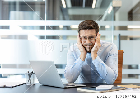 Stressed businessman experiencing an intense headache or migraine while sitting at a modern office desk, feeling overwhelmed and frustrated by work pressure and fatigue Stressed businessman experiencing an intense headache or migraine while sitting at a modern office desk, feeling overwhelmed and frustrated by work pressure and fatigue 137667462
