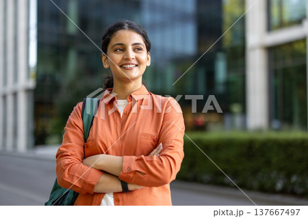 Young indian woman carrying a backpack and crossing arms, looking away and smiling in front of a modern university building, representing education, success, and future opportunities Young indian woman carrying a backpack and crossing arms, looking away and smiling in front of a modern university building, representing education, success, and future opportunities 137667497