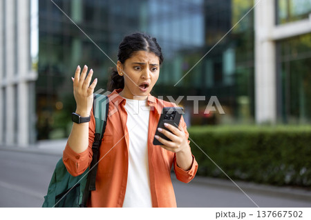 Young woman standing outdoors expresses disbelief and shock while receiving or reading bad news on her mobile phone, reflecting concepts of surprise, communication, and emotional reaction Young woman standing outdoors expresses disbelief and shock while receiving or reading bad news on her mobile phone, reflecting concepts of surprise, communication, and emotional reaction 137667502