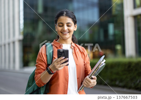 Young smiling indian woman student with backpack and notebooks checking content on her mobile phone while walking outdoors on university campus, embodying modern education and technology integration Young smiling indian woman student with backpack and notebooks checking content on her mobile phone while walking outdoors on university campus, embodying modern education and technology integration 137667503