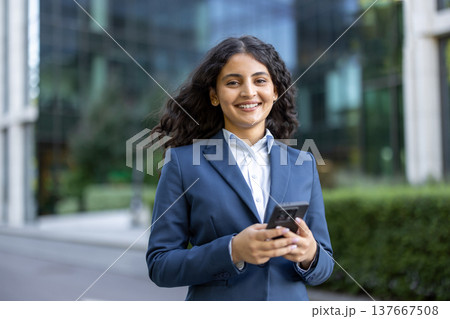 Young professional woman with dark curly hair smiles confidently while holding a smartphone outside modern office buildings, symbolizing urban career success and digital networking 137667508